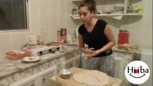 A picture of a kitchen, with Hiba Najem standing behind a table which has a piece of pita bread.
